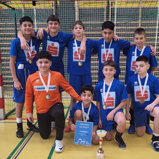 Eine Gruppe junger Jungen in blauen Trikots und Medaillen posiert für ein Foto mit einem Pokal und einem Zertifikat in einer Sporteinrichtung.