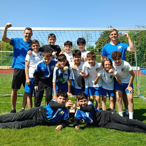 Eine Jugendfußballmannschaft posiert für ein Foto mit ihrem Pokal vor einem Fußballtor bei blauem Himmel.