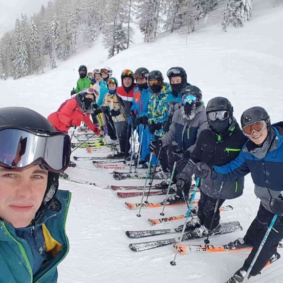Eine Gruppe von Skifahrern auf einem verschneiten Hang. Sie tragen Winterkleidung und Helme. Sie halten Skistöcke und stehen in einer Reihe. Im Hintergrund sind Kiefern zu sehen.