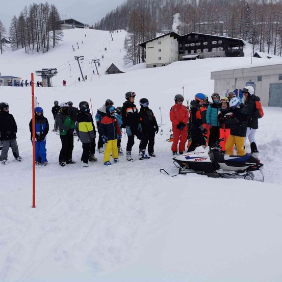 Eine Gruppe von Menschen in Winterkleidung, einschließlich Helmen und Schutzbrillen, steht in einem verschneiten Bereich in der Nähe eines Schneemobils. Ein Gebäude mit Schornstein ist im Hintergrund zu sehen.