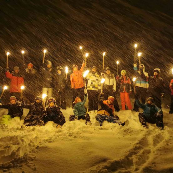 Eine Gruppe von Menschen in Winterkleidung ist im Schnee versammelt, hält brennende Fackeln, einige sitzend und einige stehend, unter einem verschneiten Himmel.