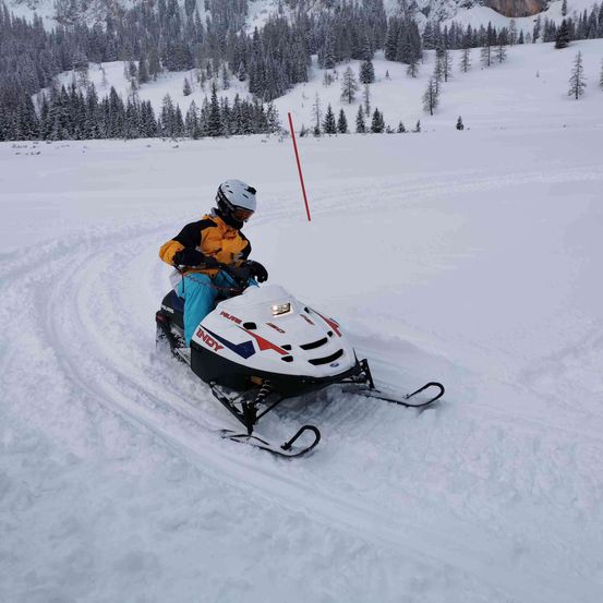Eine Person fährt mit einem Schneemobil durch eine verschneite Landschaft. Der Fahrer trägt eine gelbe Jacke, blaue Hosen und einen weißen Helm.