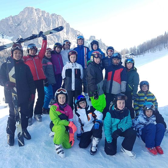 Eine Gruppe von Kindern und Jugendlichen posiert für ein Foto im Schnee. Sie tragen Wintersportausrüstung und lächeln. Verschneite Berge sind im Hintergrund.