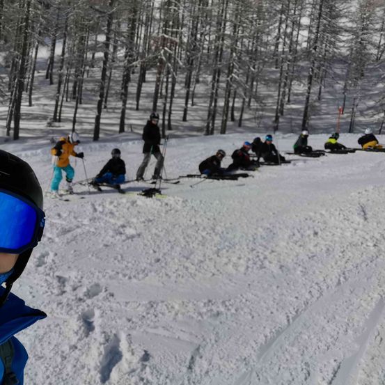 Eine Gruppe von Skifahrern und Snowboardern macht eine Pause auf einem verschneiten Berg, einige sitzen und andere stehen. Bäume umgeben das Gebiet, und ein Skifahrer im Vordergrund trägt einen Helm und eine Brille.
