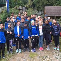 Eine Gruppe von Kindern in passenden blauen Jacken steht gemeinsam auf einem Pfad und lächelt für ein Foto. Im Hintergrund befindet sich eine kleine Hütte und eine Statue.