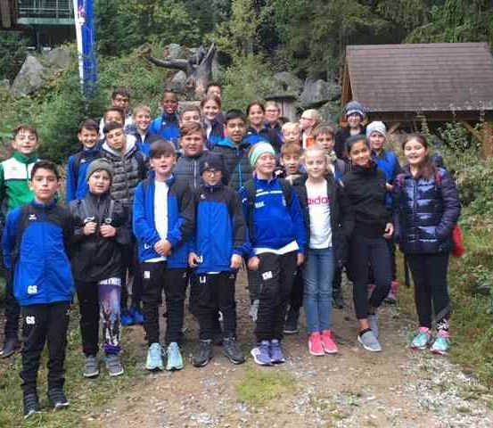 Eine Gruppe von Kindern in passenden blauen Jacken steht gemeinsam auf einem Pfad und lächelt für ein Foto. Im Hintergrund befindet sich eine kleine Hütte und eine Statue.