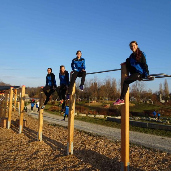 Vier Mädchen sitzen an einem sonnigen Tag auf horizontalen Stangen in einem Outdoor-Spielplatz. Sie tragen blaue Jacken und schwarze Hosen.