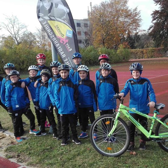 Eine Gruppe junger Radfahrer in blauen Jacken und Helmen posiert für ein Foto mit einem grünen Fahrrad auf einem Rasenfeld. Im Hintergrund befindet sich ein Banner mit dem Bild eines Radfahrers.