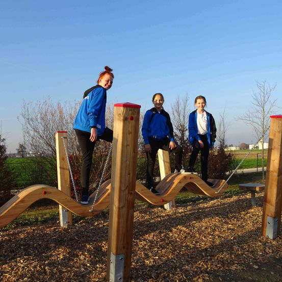 Drei Mädchen in blauen Jacken posieren auf einer wellenförmigen Holzstruktur in einem Park. Hinter ihnen befindet sich ein Feld mit Bäumen.
