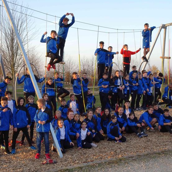 Eine Gruppe junger Menschen in blauen Uniformen posiert für ein Foto auf einem Spielplatz mit Schaukeln und einer Metallstruktur. Einige sitzen, andere stehen auf den Schaukeln.