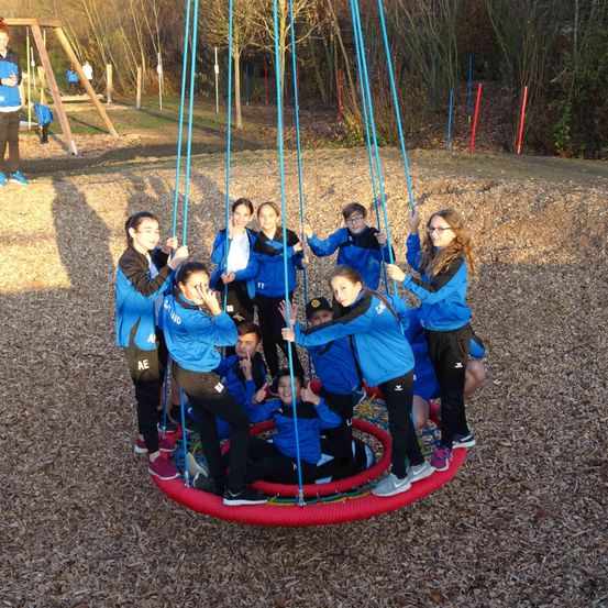 Eine Gruppe von Kindern in blauen Jacken hält sich an einer Schaukel auf einem Spielplatz fest. Der Boden ist mit kleinen, getrockneten Blättern bedeckt.