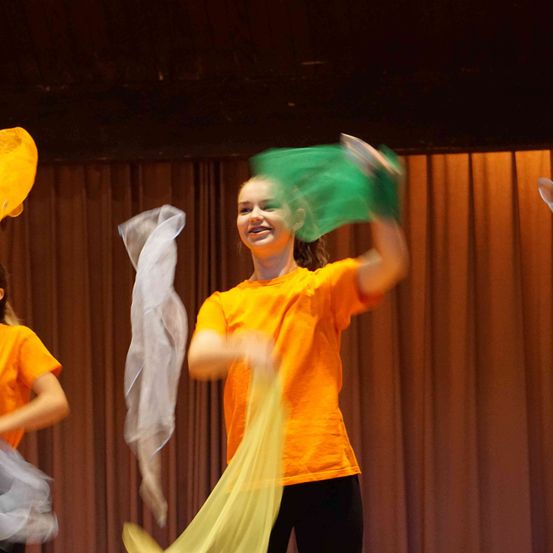 Zwei junge Mädchen in orangefarbenen Shirts führen eine Tanzaufführung auf der Bühne auf, eine hält ein grünes Tuch und die andere ein weißes.