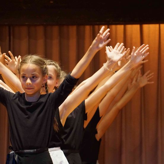 Eine Gruppe junger Tänzer tritt auf der Bühne auf. Sie tragen alle schwarze Kleidung. Das Mädchen vorne hebt beide Hände.