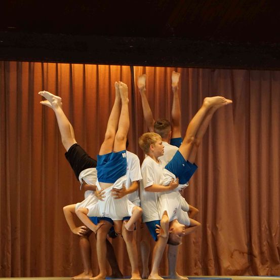 Eine Gruppe von Turnern führt eine Pyramiden-Akrobatik auf der Bühne vor, mit braunen Vorhängen im Hintergrund.