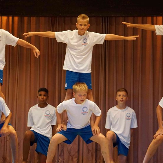 Eine Gruppe von Jungen in weißen T-Shirts und blauen Shorts führt eine Akrobatik auf der Bühne vor, mit einem braunen Vorhang im Hintergrund.