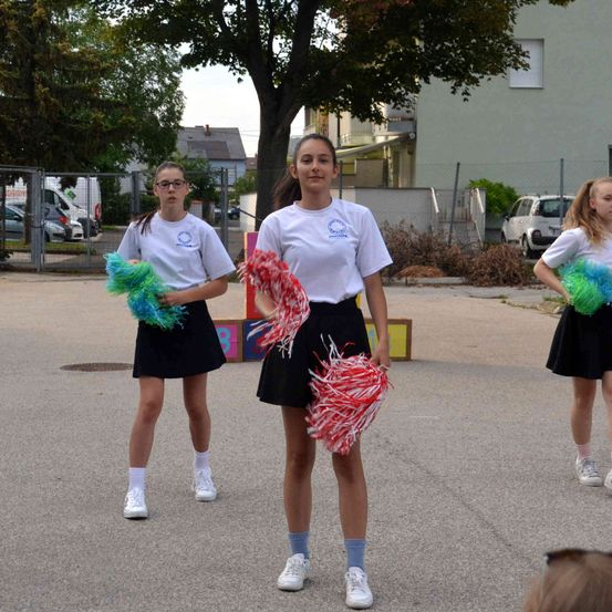 Drei junge Mädchen, die gleiche weiße Hemden und schwarze Röcke tragen, stehen auf einer Straße und halten Pom-Poms. Dahinter befinden sich Bäume, ein Zaun und Gebäude.
