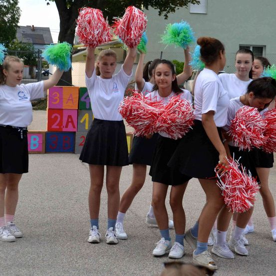 Eine Gruppe junger Mädchen, möglicherweise Cheerleader, steht in einer Reihe und hält Pom-Poms. Sie tragen weiße Hemden und schwarze Röcke. Hinter ihnen steht eine bunte Würfelstruktur.