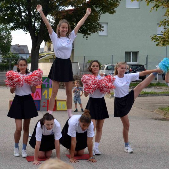 Eine Gruppe junger Mädchen in weißen und schwarzen Uniformen führt eine Cheerleader-Routine auf dem Bürgersteig durch. Eines der Mädchen steht auf den Schultern der anderen.