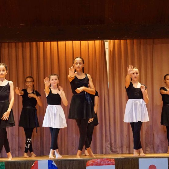 Eine Gruppe junger Mädchen in Schwarz tritt auf der Bühne auf. Sie heben die Hände und scheinen zu tanzen. Die Bühne hat einen Holzboden und einen Vorhang als Hintergrund.