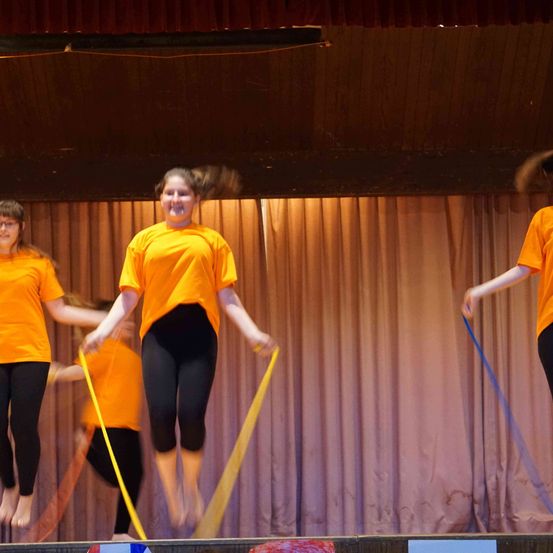 Drei Personen in orangefarbenen T-Shirts und schwarzen Hosen springen auf einer Bühne mit gelben und blauen Bändern. Im Hintergrund befindet sich ein brauner Vorhang.