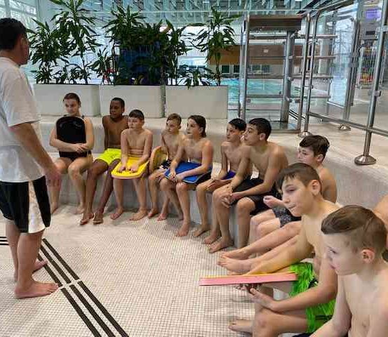 Ein Schwimmlehrer gibt einer Gruppe von Kindern, die auf einer Bank in einem Schwimmbadbereich sitzen, eine Lektion.