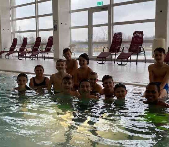 Eine Gruppe von Jungen und Mädchen ist in einem Schwimmbecken, lächelt und posiert für ein Foto. Sie sind von Glaswänden umgeben, mit Stühlen draußen. Der Raum hat eine weiße Decke und eine gelbe Rutsche auf der rechten Seite.