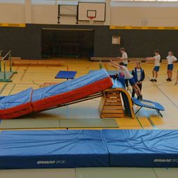 Eine Gruppe von Menschen in einer Turnhalle springt auf eine gepolsterte Rampe, während andere zusehen. Blaue Matten und ein Basketballkorb sind sichtbar.