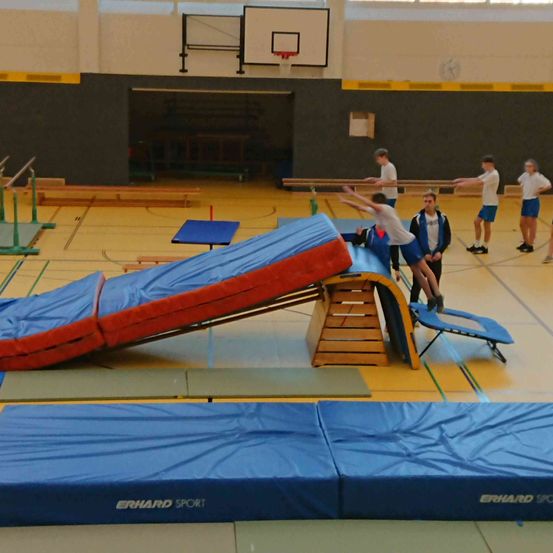 Eine Gruppe von Menschen in einer Turnhalle springt auf eine gepolsterte Rampe, während andere zusehen. Blaue Matten und ein Basketballkorb sind sichtbar.