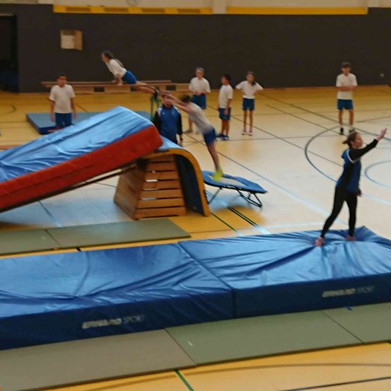 Mehrere Kinder üben Gymnastik in einer Turnhalle, einige auf blauen Matten und andere beobachten. Ein Kind springt auf eine blaue Matte.