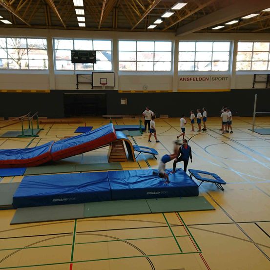 Sporthalle mit Gymnastikmatten, einer Rutsche und einem Basketballkorb. Mehrere Personen üben Akrobatik und eine springt auf einer Matte.