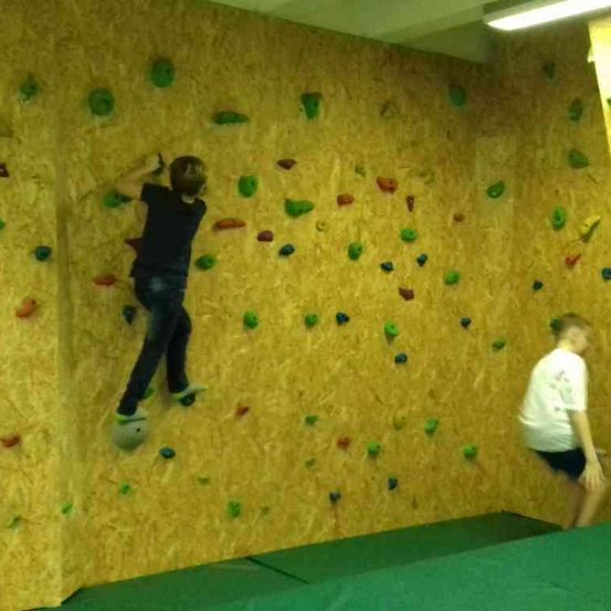 Zwei Jungen klettern an einer Wand mit bunten Griffen. Ein Junge in Schwarz klettert höher. Ein anderer Junge in Weiß steht an der Basis der Wand.