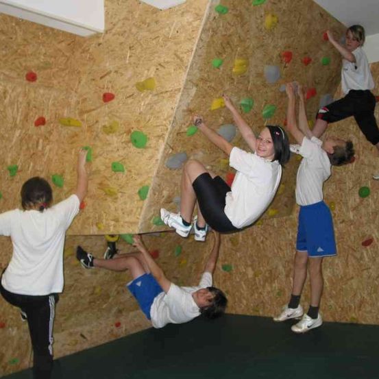 Eine Gruppe von Kindern klettert an einer Wand mit verschiedenen farbigen Griffen. Jedes Kind trägt weiße T-Shirts und Turnschuhe. Ein Kind hängt kopfüber, während ein anderes an der Wand klettert.