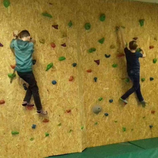 Zwei Kinder klettern an einer Holzwand mit verschiedenen farbigen Griffen. Ein Kind trägt ein grünes Shirt und schwarze Hose, das andere ein dunkles Shirt und dunkle Hose.