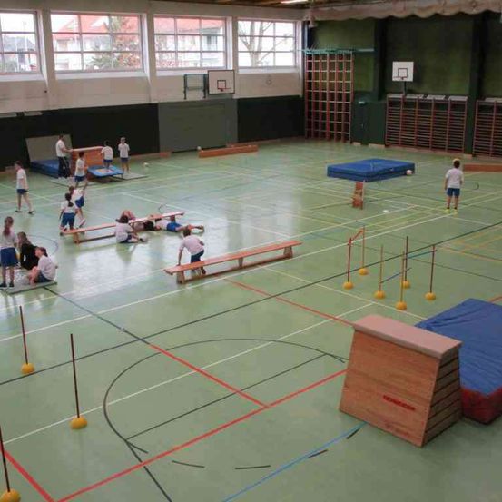 Eine Sporthalle mit grünem Boden hat mehrere spielende Kinder. Einige liegen auf Bänken, andere stehen. Es gibt blaue Matten, Holzhindernisse und einen Stapel Holzkisten.