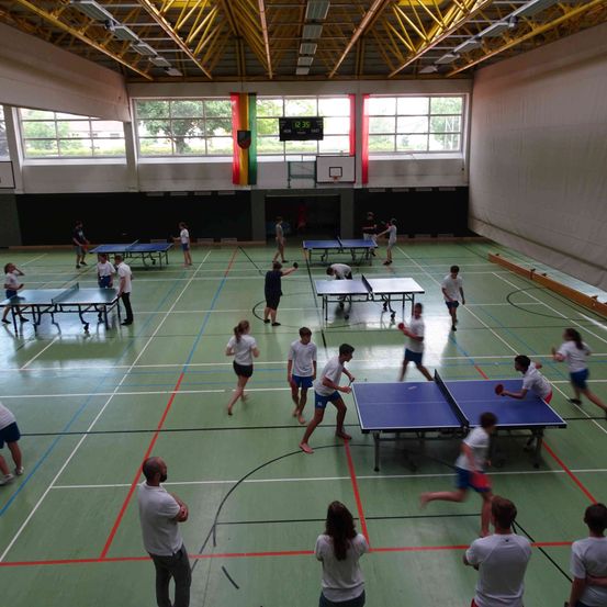 Im Inneren, mehrere Tische, Spieler beim Tischtennis, Zuschauer beobachten, Flaggen und Anzeigetafel sichtbar.