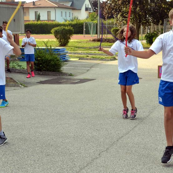 Eine Gruppe von Kindern spielt mit Sprungseilen auf einem Schulhof. Einige tragen Turnschuhe, und einer hält ein rotes Seil. Dahinter befinden sich Büsche und ein Zaun.