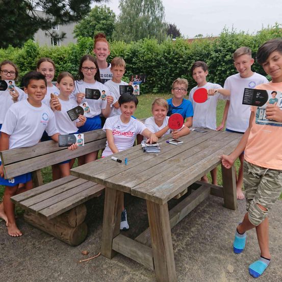 Eine Gruppe junger Kinder posiert für ein Foto mit Schlägern, Büchern und Karten, die auf einer Holzbank mit einem Tisch sitzen. Sie scheinen sich in einem Park zu befinden.