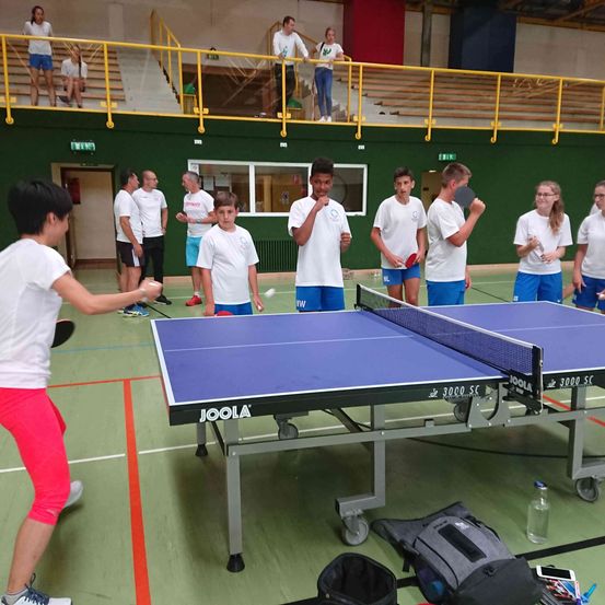 Eine Gruppe junger Menschen spielt Tischtennis auf einem Platz mit einem Joola-Tisch. Mehrere Personen schauen von den Tribünen zu.