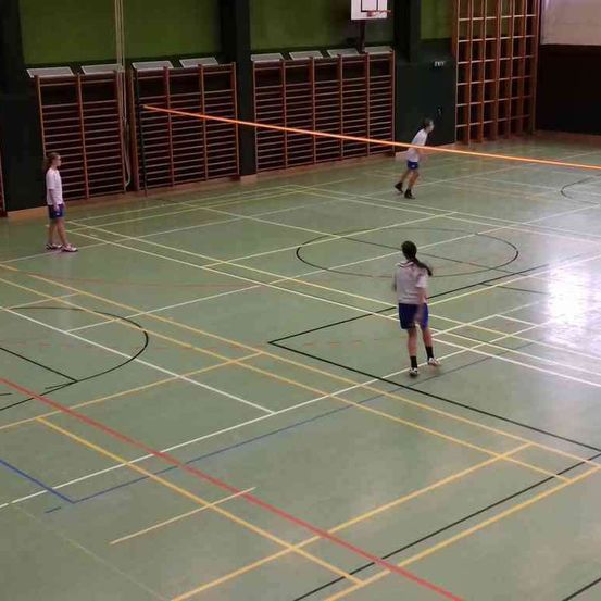 Drei Personen spielen Badminton in einer Halle. Ein Spieler hält einen Schläger, während die anderen beiden an gegenüberliegenden Enden stehen. Der Platz hat Linien und Markierungen auf dem Boden.