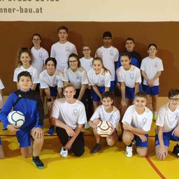 Eine Gruppe junger Athleten in weißen und blauen Uniformen posiert mit ihren Fußballbällen für ein Foto in einer Turnhalle. Die Wand hinter ihnen hat eine Website-Adresse.