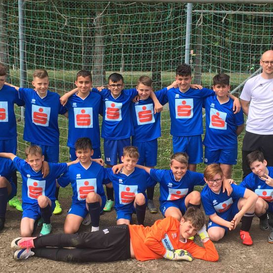 Eine Jugendfußballmannschaft posiert für ein Foto vor einem Netz. Sie tragen blaue Trikots mit einem Logo, außer einem, der in Orange liegt. Hinter ihnen steht ein Mann mit Brille und einem weißen T-Shirt.