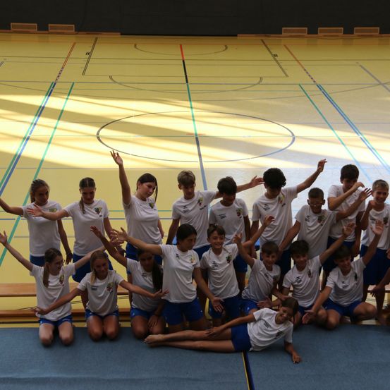 Eine Gruppe junger Athleten in weißen und blauen Uniformen posiert für ein Foto auf einem Sportplatz mit gelbem Boden und blauen Markierungen.