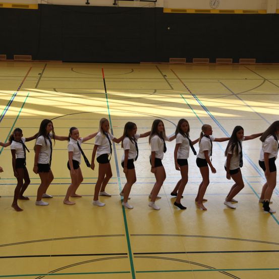 Eine Gruppe junger Mädchen in weißen Shirts und schwarzen Shorts übt eine koordinierte Tanzroutine auf einem Hallenplatz.