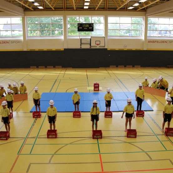Eine Gruppe von Kindern in gelben Uniformen und weißen Helmen steht auf roten Kisten in einer Turnhalle. Eine blaue Matte liegt auf dem Boden.