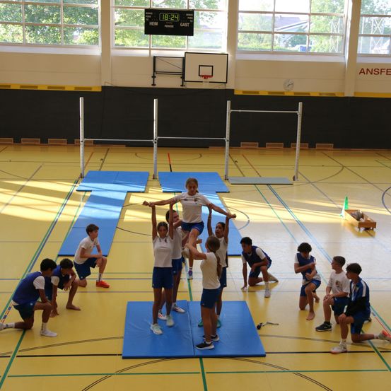 Eine Gruppe von Kindern in einer Turnhalle, die Gymnastik auf blauen Matten übt, mit einer Anzeigetafel im Hintergrund.