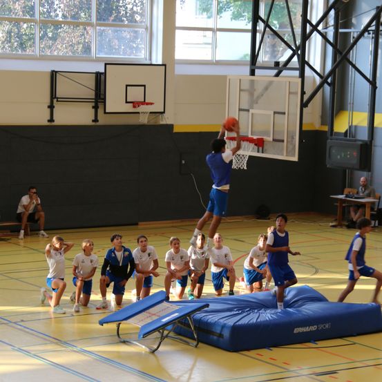 Eine Gruppe junger Basketballspieler trainiert in einer Turnhalle, ein Spieler dunkt den Ball, während andere zuschauen und ein Trainer von der Seite beobachtet.