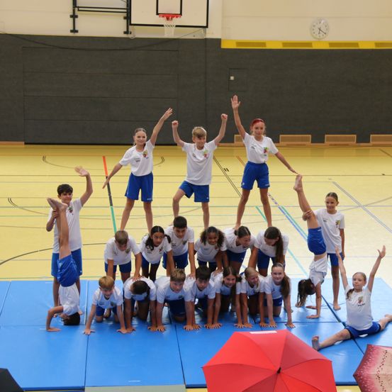 Eine Gruppe von Kindern in weißen T-Shirts und blauen Shorts bildet eine Pyramide auf einem blauen Teppich in einer Turnhalle. Einige Kinder stehen auf den Schultern der anderen.