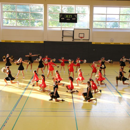 Eine Gruppe Cheerleader in roten Uniformen führt auf einem Basketballplatz eine Darbietung durch. Der Platz hat gelbe Markierungen und eine Anzeigetafel zeigt die Zeit an.
