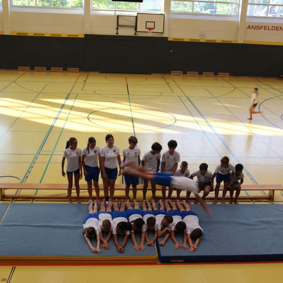 Eine Gruppe von Kindern in blauen und weißen Uniformen führt Akrobatik auf einem blauen Matten in einer Turnhalle aus. Ein Junge macht einen Handstand, während andere zusehen.