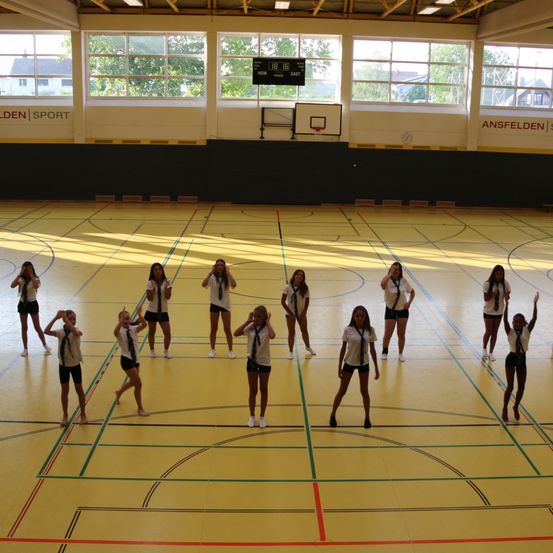 Eine Gruppe junger Mädchen in einheitlichen Kostümen führt eine synchronisierte Tanzroutine auf einem überdachten Basketballplatz auf. Der Platz ist hell beleuchtet und die Wände sind mit Werbung geschmückt.